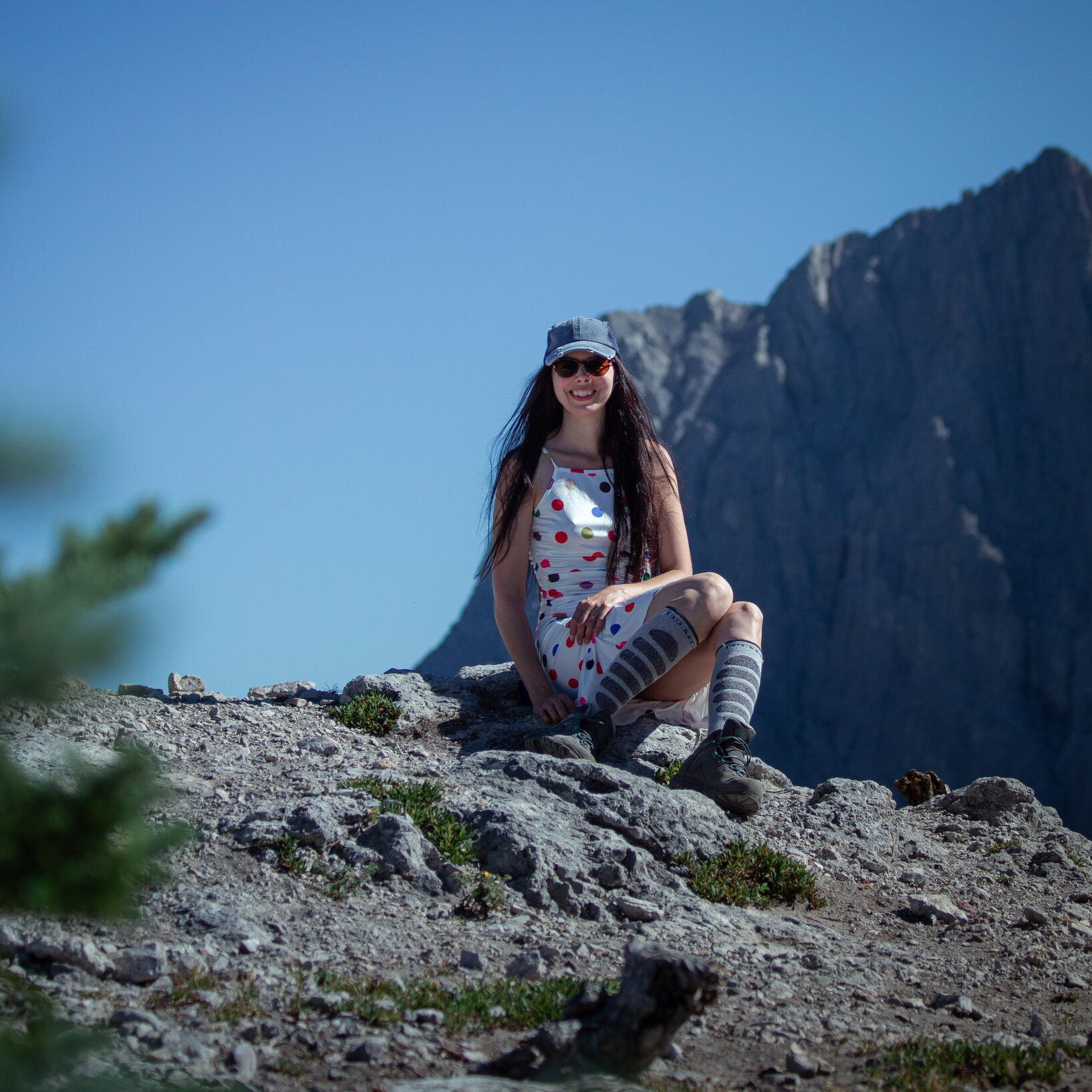 Layla Messner in a polkadot dress by David Dethlefs A happy brunette in a polkadot dress, a denim ball cap, and hiking boots sitting on a rock in the alpine.